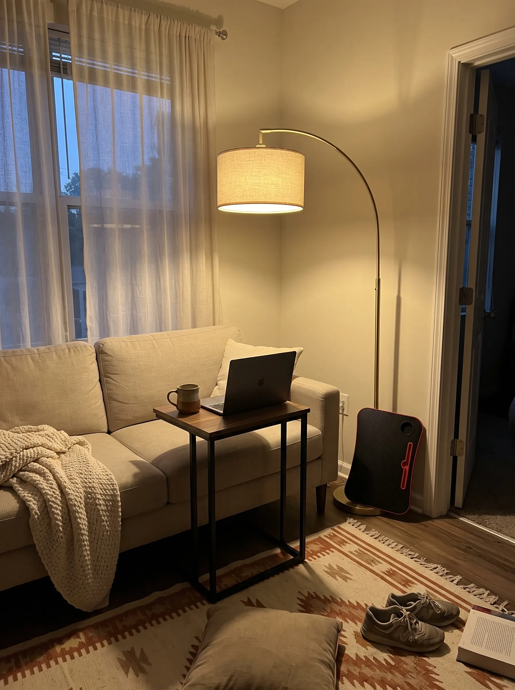 College apartment living room with a C-shaped side table beside a couch, an arc floor lamp casting warm light, and a foldable lap desk, college apartment must haves for a multipurpose room